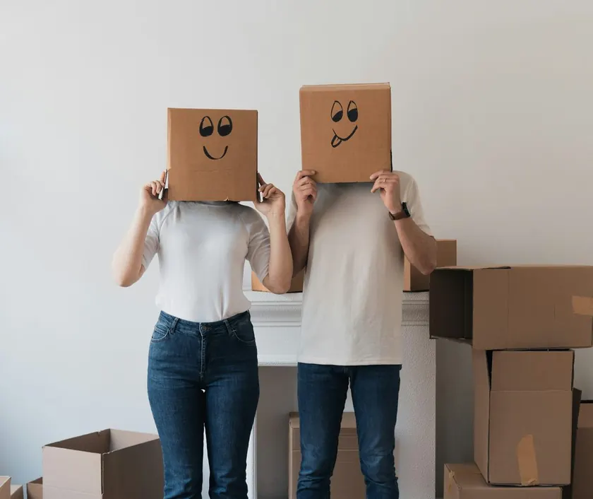 A couple holding moving boxes in front of their faces in their new home