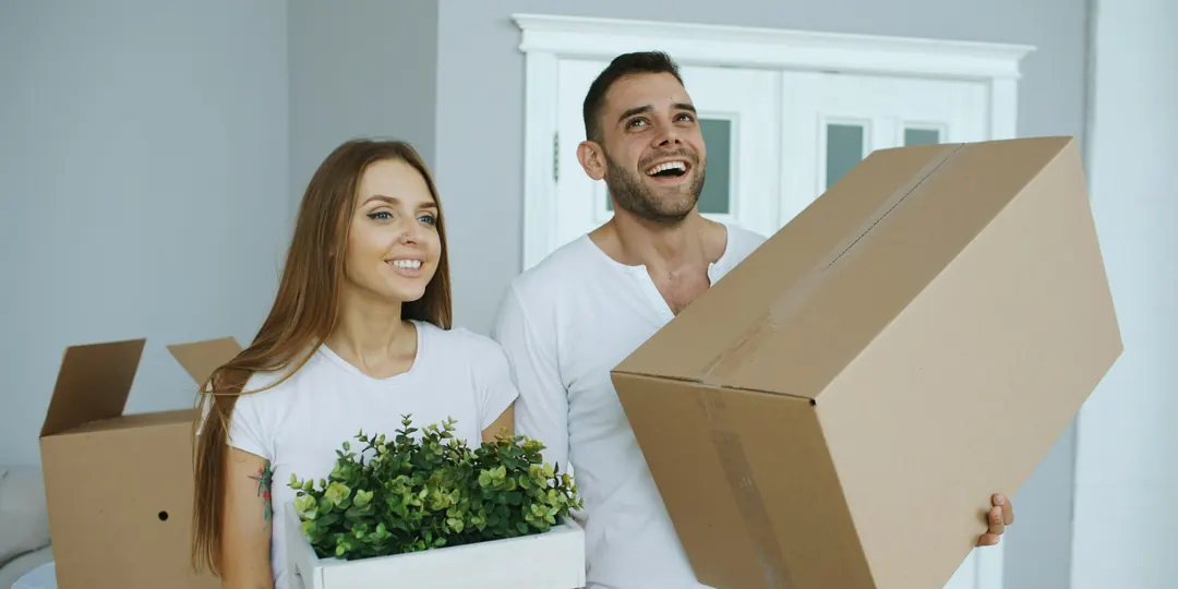 A smiling couple carrying boxes and a plant into their first home